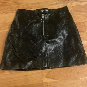 Svart kort kjol i skinnimitation - Snygg svart kort kjol i glansig skinnimitation med dragkedja framtill och två silverfärgade knappar i midjan. Kjolen har en rak passform