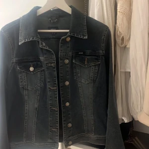 Svart jeansjacka från Mexx, stl 38 - Snygg svart jeansjacka från Mexx i klassisk modell med silverfärgade knappar och två bröstfickor. Jackan har markerade sömmar och en cool, mörk tvätt som passar perfekt till alla outfits. Stängs med knappar framtill och har lång ärm.