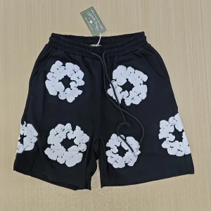 Svarta shorts från Universal Overall - Svarta shorts med vita blommiga tryck från Denim Tears. De har elastisk midja med snörning och praktiska fickor. Perfekta för en chill sommarlook med sin loose passform och coola print.