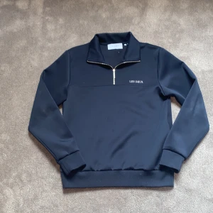 Svart half zip tröja från Les Deux - Svart långärmad tröja från Les Deux med half zip och stilren vit logga på bröstet. Tröjan har hög krage och är gjord i mjukt material som känns skönt mot huden. Perfekt för dig som gillar en clean och enkel look. Hör av dig vid minsta fundering så svarar jag direkt! Priset är prutbart.
