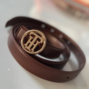 Brunt skärp från Tommy Hilfiger - Snyggt brunt bälte från Tommy Hilfiger med rund, guldig metallspänne och TH-logga framtill. Bältet är gjort i slätt skinn och har flera hål för justering. Perfekt accessoar för att lyfta din outfit med en stilren detalj.