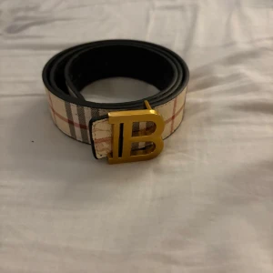 Burberry rutig bälte med guldspänne - Säljer ett snyggt Burberry bälte med klassiskt rutigt mönster i beige, svart och rött. Bältet har ett stort guldigt B-spänne framtill och är tillverkat i skinn. Perfekt för att lyfta din outfit med en lyxig touch.