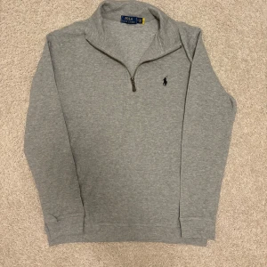 Grå half zip tröja Polo Ralph Lauren - Snygg grå långärmad tröja från Polo Ralph Lauren med klassisk half zip-dragkedja och broderad logga på bröstet. Skön passform och perfekt för lager-på-lager. Tillverkad i mjuk bomull för en chill och stilren look.