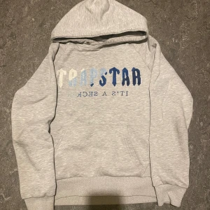 Grå Trapstar hoodie med blå text - Säljer en grå hoodie från Trapstar i storlek S men passar dig som är 1,65 - 1,73. Tröjan har tryck framtill med texten 'TRAPSTAR' i olika blå nyanser och 'IT'S A SECRET' under. Klassisk känguruficka och huva, perfekt för chill dagar. Mjuk och skön bomullsblandning.