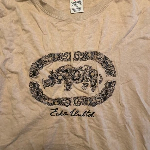 Beige t-shirt från Ecko Unltd. - Snygg beige t-shirt från Ecko Unltd. med stort svart tryck på bröstet som har detaljerade mönster och döskallar. Rund hals och klassisk passform. Tillverkad i 100% bomull för skön känsla. Perfekt för dig som gillar streetwear och coola prints.