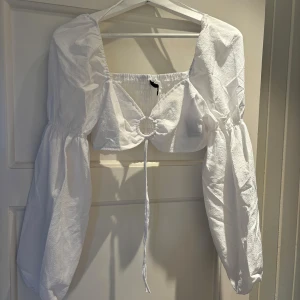 Vit croppad blus med puffärmar från H&M - Superfin vit croppad blus med långa puffärmar och fyrkantig ringning. Toppen har en snygg ringdetalj framtill med knytband och smockad rygg för extra komfort. Perfekt för dig som vill sticka ut med en trendig och luftig look.