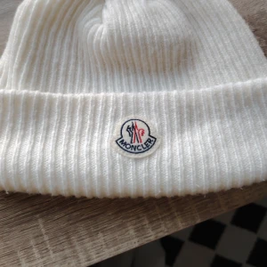 Vit ribbstickad mössa från Moncler - Vit ribbstickad mössa från Moncler med klassisk uppvikt kant och broderad logotyp framtill. Mössan är i mjukt material och har en clean, stilren look som passar perfekt till kalla dagar.