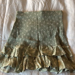 Grön volangkjol från Mayoral - Supersöt grön kortkjol från Mayoral med smockad midja och dubbla volanger nertill. Kjolen har ett diskret blommönster i vitt och känns lätt och luftig, perfekt för varmare dagar. Snygg att matcha med en enkel topp eller stickad tröja.