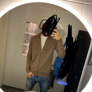 Beige kofta - Säljer en stilren beige kofta med hel dragkedja framtill. Tröjan är stickad och har långärmad design med ribbade muddar och nederkant. Perfekt för lager-på-lager och passar till jeans eller chinos. Hör av dig vid fler funderingar!
