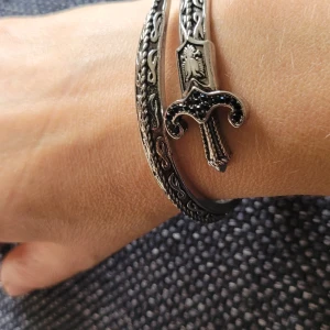  Helt ny Silvrigt armband med svärd-detalj -  Helt ny Unikt armband i silverfärgad metall med coolt svärd som detalj framtill. Armbandet har ingraverade mönster längs hela ytan och svärdets handtag är dekorerat med svarta stenar. Perfekt accessoar för dig som gillar edgy och annorlunda smycken.passar till en kvinna eller kille 