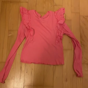 Rosa volangtröja från Kappahl - Supersöt rosa långärmad topp från Kappahl med volangdetaljer längs axlarna. Toppen är ribbad och har en rund halsringning. Perfekt för dig som gillar färg och vill sticka ut lite extra.