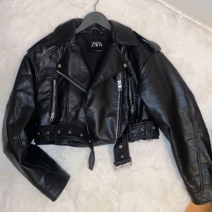 Svart skinnjacka från Zara med bälte - Snygg svart skinnjacka från Zara i bikerstil med silvriga dragkedjor, bred krage och bälte i midjan. Jackan är croppad och har coola detaljer som axelklaffar och flera fickor med dragkedja. Perfekt för dig som vill ha en edgy look.