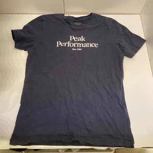 Mörkblå Peak Performance t-shirt - Snygg mörkblå t-shirt från Peak Performance med vit logga och text på bröstet. Klassisk rund halsringning och korta ärmar. Perfekt för en chill och sportig look. Gjord i mjuk bomull som känns skön mot huden. Mvh Hannes!
