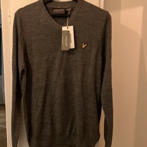 Grå v-ringad tröja från Lyle & Scott - Snygg grå v-ringad tröja från Lyle & Scott med klassisk gul logga på bröstet. Tröjan är långärmad och har en stilren, enkel design som passar perfekt till jeans eller chinos. Tillverkad i mjukt material som känns skönt mot huden.