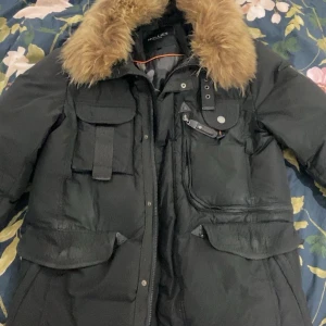 Svart dunjacka med fuskpäls från Hollies - Säljer en svart dunjacka från Hollies med stor brun fuskpäls runt kragen. Jackan har flera fickor med lock och dragkedja, samt camouflagemönstrat innerfoder. Perfekt för kalla dagar och har en cool, urban vibe.