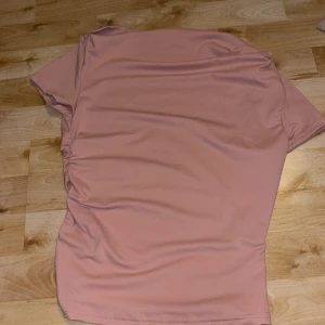 Rosa off shoulder topp med rynkade sidor - Säljer en ljusrosa topp med korta ärmar och snygga rynkade detaljer längs ena sidan. Toppen har en enkel och stilren design i mjukt syntetmaterial, perfekt att matcha med jeans eller kjol.