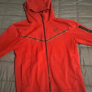 Röd Nike hoodie med dragkedja M - Nike hoodie i rött med hel dragkedja och svart logga på bröstet. Tröjan har huva, långa ärmar och en snygg svart detalj på ena ärmen. Perfekt för dig som gillar sportig och stilren look.