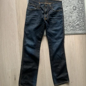 Mörkblå jeans från Nudie Jeans, 34/34 - Säljer ett par mörkblå jeans från Nudie Jeans i klassisk rak modell. Jeansen har kontrastsömmar i orange, fem fickor och läderpatch med logga bak. Tillverkade i slitstark denim av bomull. Perfekta för dig som gillar stilrena och tidlösa jeans.