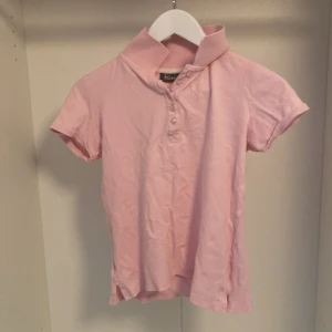 Ljusrosa pikétröja från Mira - Säljer en ljusrosa pikétröja från Mira i klassisk modell med krage och knappar framtill. T-shirten är kortärmad och tillverkad i mjuk bomull, perfekt för en chill och stilren look. Passar dig som gillar enkel och fräsch stil.