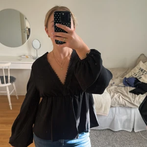 Svart blus med spets och v-ringning - Säljer en svart blus med djup v-ringning och fina spetsdetaljer längs halsringningen. Blusen har långa, puffiga ärmar och en lös passform under bysten. Perfekt för dig som gillar en fin och trendig stil💓