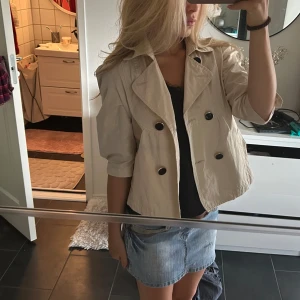 Beige kort trenchcoat  - Snygg beige kort trenchcoat med bred krage och stora svarta knappar framtill. Jackan har trekvartsärmar och en avslappnad passform. Perfekt för lager-på-lager och ger en cool vibe till din outfit.
