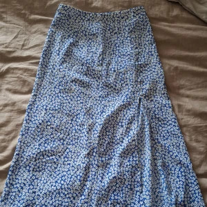 Blåvit blommig långkjol med slits - Snygg långkjol i blått med vita små blommor och en diskret slits på sidan. Kjolen har en smickrande A-linjeform och stängs med dragkedja i sidan. Perfekt för dig som gillar ett feminint och somrigt uttryck.