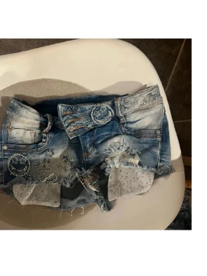 Blå jeansshorts med pärlor och paljetter - Unika blå jeansshorts med slitna detaljer, pärlor och paljetter på framsidan. Shortsens kanter är fransiga och de har dekorativa bälteshällor samt genomskinliga tygdetaljer med glitter. Perfekta för dig som vill sticka ut i sommar! Midjemått 36