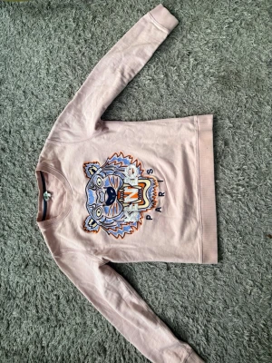 Rosa sweatshirt med tiger från Kenzo - Säljer en ljusrosa sweatshirt från Kenzo med ikoniskt broderat tigerhuvud och texten 'Paris' på bröstet. Tröjan har rund hals, långa ärmar och är gjord i mjuk bomull. Perfekt för dig som vill sticka ut med en klassisk streetwear-look.