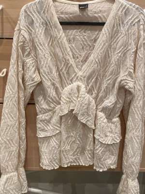 Krämvit strukturerad blus Gina Tricot - Säljer en krämvit blus från Gina Tricot med djup v-ringning, volangdetalj framtill och långärmad design. Blusen har ett strukturerat, våfflat tyg och breda muddar vid ärmsluten. Perfekt för dig som gillar trendiga detaljer och en unik look.