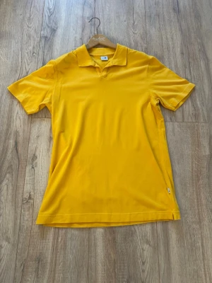 Gul piké t-shirt från NN07 - En stilren gul piké t-shirt med klassisk krage och korta ärmar. Tillverkad i mjuk bomull som känns skön mot huden. Enkel design utan mönster eller tryck, perfekt för dig som gillar färg och vill ha en clean look. I nyskick.