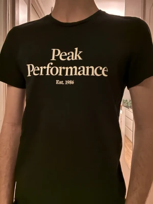Svart Peak Performance t-shirt - Snygg svart t-shirt från Peak Performance med vit logga och texten 'Est. 1986' på bröstet. Klassisk rund halsringning och korta ärmar. Perfekt för en clean och stilren look.