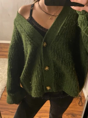 Grön oversized kofta med guldknappar - Mysig grön kofta i oversized modell med stora guldknappar framtill. Koftan har en djup v-ringning och är gjord i ett fluffigt, stickat material. Perfekt att slänga över en topp för en chill look.