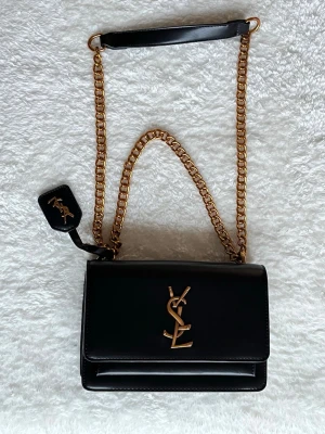 Svart YSL handväska i skinn - Snygg svart handväska från Saint Laurent med guldfärgad kedjerem och ikoniskt YSL-emblem framtill. Väskan är tillverkad i slätt skinn och har en stilren, rektangulär form. Perfekt för dig som vill ha en klassisk och lyxig accessoar.