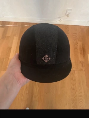 Svart ridhjälm från Charles Owen - Säljer en klassisk svart ridhjälm från Charles Owen med sammetsliknande yta och en diskret logga framtill. Hjälmen har en rundad form och en kort skärm. Perfekt för dig som vill ha en stilren look i stallet.