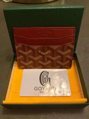 Röd kortplånbok från G - Snygg och exklusiv kortplånbok från G i rött med klassiskt mönster och logga framtill. Plånboken har flera kortfack och är tillverkad i skinn med detaljer i brunt och beige. Perfekt för dig som vill ha stilren och lyxig accessoar.