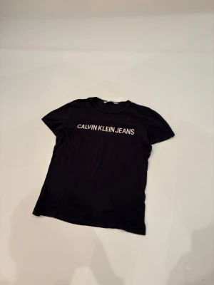  Calvin Klein T-shirt - Svart t-shirt från Calvin Klein Jeans med klassisk rund hals och vit logotyptryck över bröstet. Enkel och stilren design som passar perfekt till jeans eller shorts. Tillverkad i mjuk bomull för en bekväm känsla hela dagen.