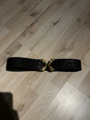 Unikt vintagebälte 1980-tal – statement spänne med djurmotiv - Unikt vintagebälte från 1980-talet i svart präglat äkta läder med ett tungt, dekorativt guldfärgat statement-spänne med djurhuvuden.  Typisk kraftfull 80-talsdesign och ett riktigt blickfång. Perfekt att bära över kavaj, klänning eller ytterplagg för en markerad silhuett.  🔹 Äkta läder 🔹 Bra vintage-skick med naturlig patina 🔹 Original från 80-talet 🔹 Statement-accessoar / samlarobjekt  Märkning har nötts bort med ålder (vanligt på äldre vintage), se bilder för detaljer. Hör gärna av dig!