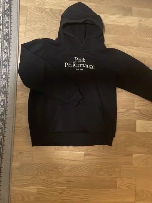 Svart hoodie från Peak Performance - Snygg svart hoodie från Peak Performance med vit logga och text på bröstet. Klassisk modell med huva och magficka, perfekt för en avslappnad stil. Mjuk bomullskänsla och ribbade muddar vid ärmslut och nederkant. Storlek s/170cm