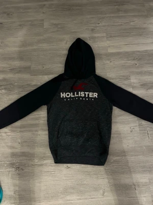 Mörkgrå Hollister hoodie med tryck - Snygg mörkgrå hoodie från Hollister med svart huva och ärmar. Stor logga och text 'HOLLISTER CALIFORNIA' i vitt och rött framtill. Klassisk känguruficka och mjukt material, perfekt för chill dagar. Passar dig som gillar streetwear och enkel stil.