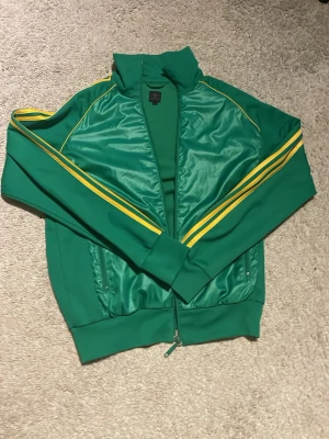 Adidas Montréal 1976 - Retro grön vindjacka från Adidas med gula ränder längs ärmarna och texten 'Montréal 1976' på ryggen. Jackan har dragkedja framtill, ribbade muddar och klassisk sportig look. Perfekt för dig som gillar vintage och streetstyle.
