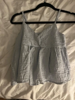 Ljusblå omlott linnetopp - Säljer en ljusblå omlott linnetopp med tunna axelband och luftig, vågig struktur. Toppen har en avslappnad passform och är perfekt för varma dagar. Materialet känns mjukt och svalt mot huden.