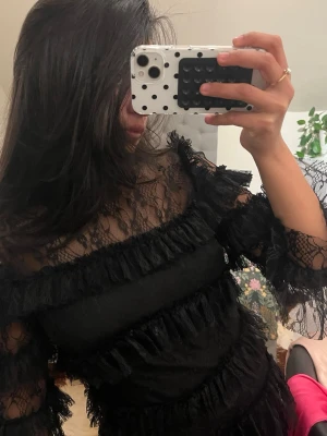 Svart spetsklänning från By Malina  - Säljer en svart klänning med transparent spets upptill och på ärmarna. Klänningen har volangdetaljer och är långärmad med en elegant, festlig känsla. Perfekt för dig som vill sticka ut med en unik och trendig look.