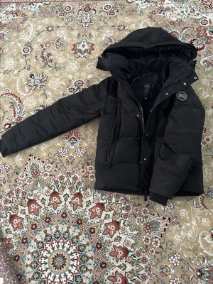 Svart pufferjacka.  Canada goose  - Svart Canada goose jacka från Canada goose. Med logga på ärmen. Jackan har dragkedja och tryckknappar framtill samt 2 fickor. Perfekt för kalla dagar och ger en modern, urban look. 