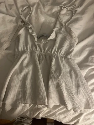 Vit linnetopp med volang från lager 157 - Supersöt vit linnetopp från 157 med tunna axelband som knyts och volangdetalj vid ringningen. Toppen har en lätt rynkad struktur och ärmlös design, perfekt för varma dagar. Passar dig som gillar en luftig och feminin stil.