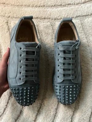 Gråa Louboutin sneakers med nitar - Säljer ett par gråa sneakers från Christian Louboutin i mocka med ikoniska röda sulor och coola nitar på tån. Skorna har snörning och en rund tå, perfekt för dig som vill sticka ut med din stil.