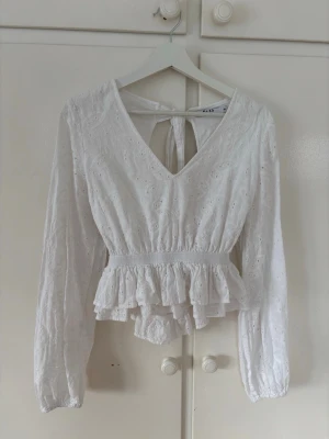 Vit blus från NA-KD - Söt vit blus från NA-KD med broderade detaljer och volang nedtill. Blusen har v-ringning, långa ärmar och öppen i ryggen med knytbara band. Storlek 38🩷