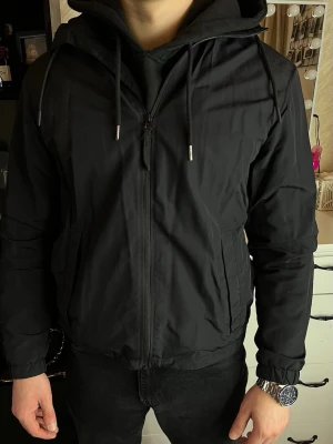 Svart vindjacka med huva - Snygg svart vindjacka från H&M med huva och dragsko. Helt som nya. Jacka i storlek S men passar även M (jag brukar har storlek M). Ser lite skrynklig ut eftersom den har legat i garderoben.