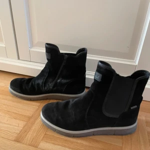 Svarta kängor i mocka storlek 39 Goretex  - Snygga svarta kängor i mocka med rund tå och dragkedja på sidan. Skorna har en platt sula och diskreta detaljer, perfekta för en stilren look. Passar till många olika outfits och är både bekväma och trendiga. Storlek 39 