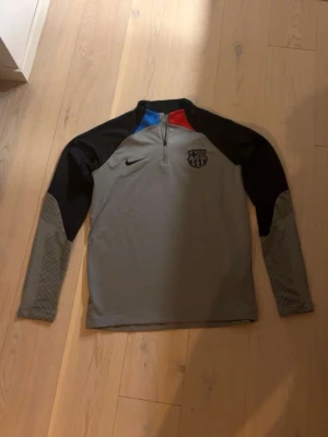 Nike FC Barcelona långärmad träningströja - Snygg långärmad träningströja från Nike med FC Barcelona-logga på bröstet. Tröjan är grå med svarta ärmar och har detaljer i blått och rött vid halsen. Materialet känns som polyester och den har en half zip framtill. Perfekt för dig som gillar fotboll och vill ha en sportig look.