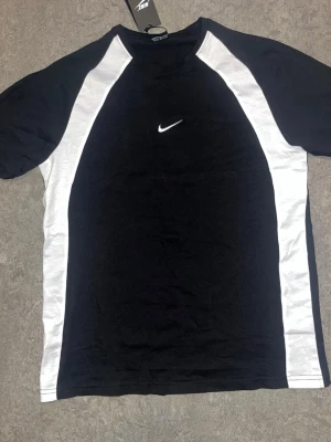 Svart och vit Nike t-shirt - Snygg svart t-shirt från Nike med vita paneler längs sidorna och ärmarna. Klassisk Nike-logga broderad på bröstet. T-shirten har korta ärmar och rund hals. Perfekt för en sportig och avslappnad stil.